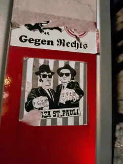 Street sticker A sticker depicting two men in suits, reminiscent of the Blues Brothers, with knuckles showing FCSP and 1910. The background features vertical stripes and the text "Forza St. Pauli".  There is also another sticker above it with the text "Gegen Rechts" visible.