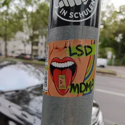 A sticker featuring a pop art style illustration of a woman's mouth with her tongue sticking out. The tongue has an LSD and MDMA stamp on it. The sticker has a QR code in the corner.