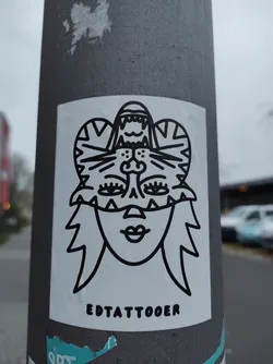 Street sticker Black and white illustration of a face wearing a tiger mask. The style is simple and bold, with sharp lines and a focus on graphic design. The sticker features the text "EDTATTOER" at the bottom, likely indicating the artist or brand.