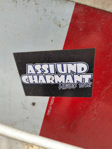 A black sticker with white and blue text that says "ASSLUND CHARMANT" and "#HIBSC 1892" in a stylized font. The sticker is placed on a white and red surface.