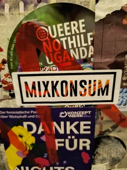 Street sticker A sticker with the word "Mixkonsum" written in bold, uppercase letters. The text is stylized with a pattern resembling tiger stripes or a similar design in orange and black, set against a white background within a black border.  The sticker appears to be placed on a wall among other stickers and posters.