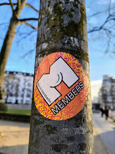 A round sticker with an orange and yellow pixelated border.  The central design features a stylized 'M' in white against an orange background. The word 'MEMBERS' is written in bold, uppercase letters.  The text '@membersberlin' is visible in smaller font at the side. The sticker is affixed to a tree trunk.