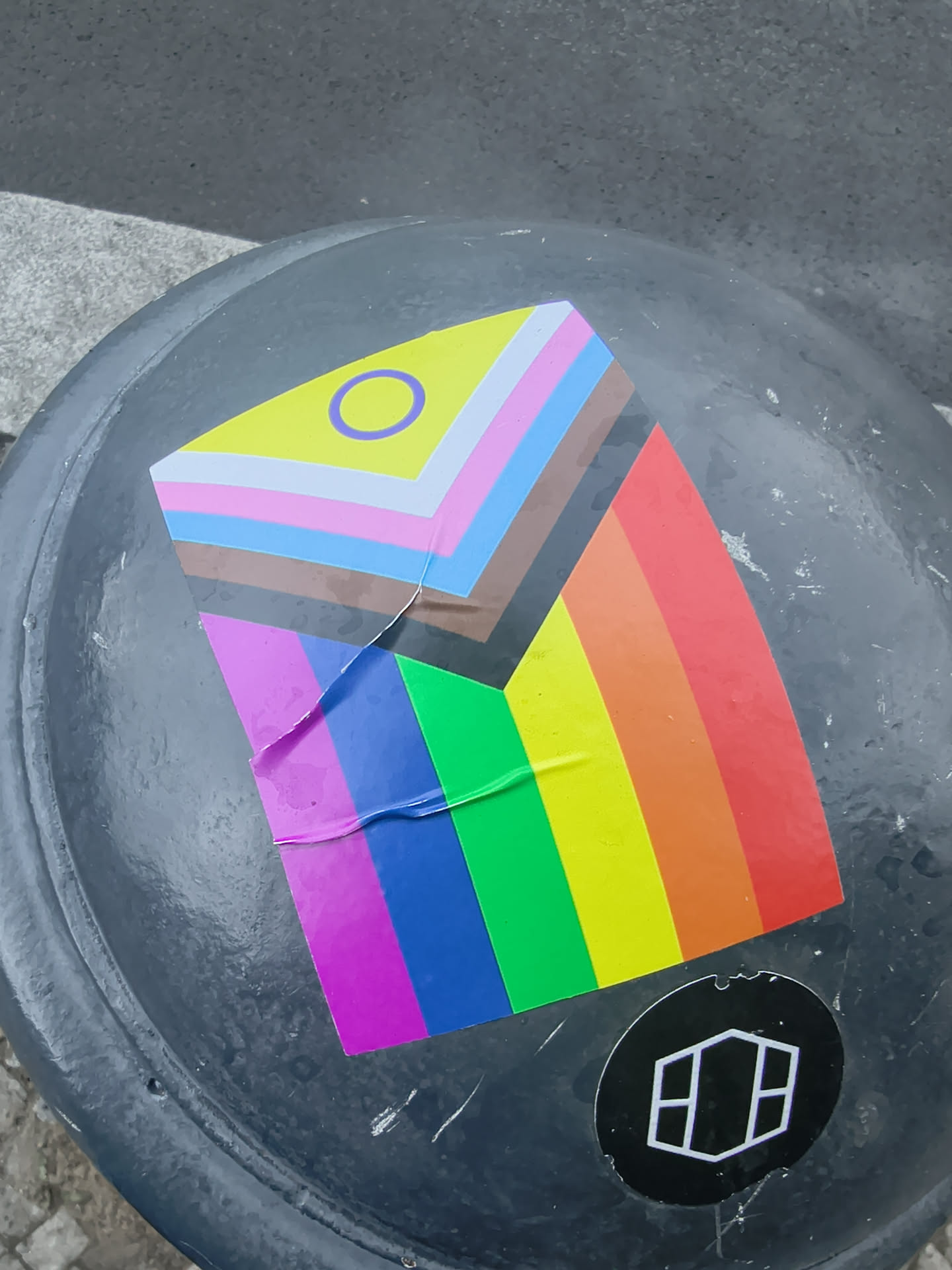 A rectangular sticker depicting the Progress Pride Flag with intersex inclusion (yellow triangle and purple circle). It is adhered to a dark, curved surface and shows slight creasing.