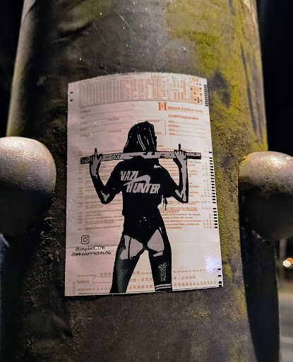 A sticker featuring a stylized female character carrying a long object across her shoulders. The character is wearing a black top with the words 'Vaz Hunter' printed on it and black bottoms. The sticker is placed on a weathered pillar. An Instagram handle is visible at the bottom. 