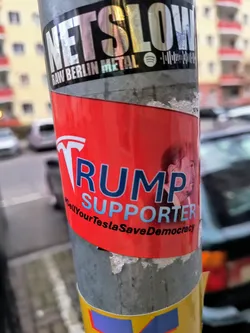 Street sticker A red sticker with the text "TRUMP SUPPORTER" and a small image of Donald Trump.  The bottom of the sticker has smaller text that appears to say "SellYourTeslaSaveDemocracy".
