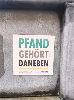 Street sticker The sticker is square-shaped with a white background. It features large, bold text in the center. The top line reads 'PFAND' in a teal color. The second line reads 'GEHÖRT' in a dark gray color. The third line reads 'DANEBEN' in a dark gray color. Below the main text, there is a thin yellow line. At the bottom left, there is a website address 'pfand-gehört-daneben.de'. To the right of the website, it says 'Eine Initiative von:' followed by the 'fritz-kola' logo in white with a black outline.