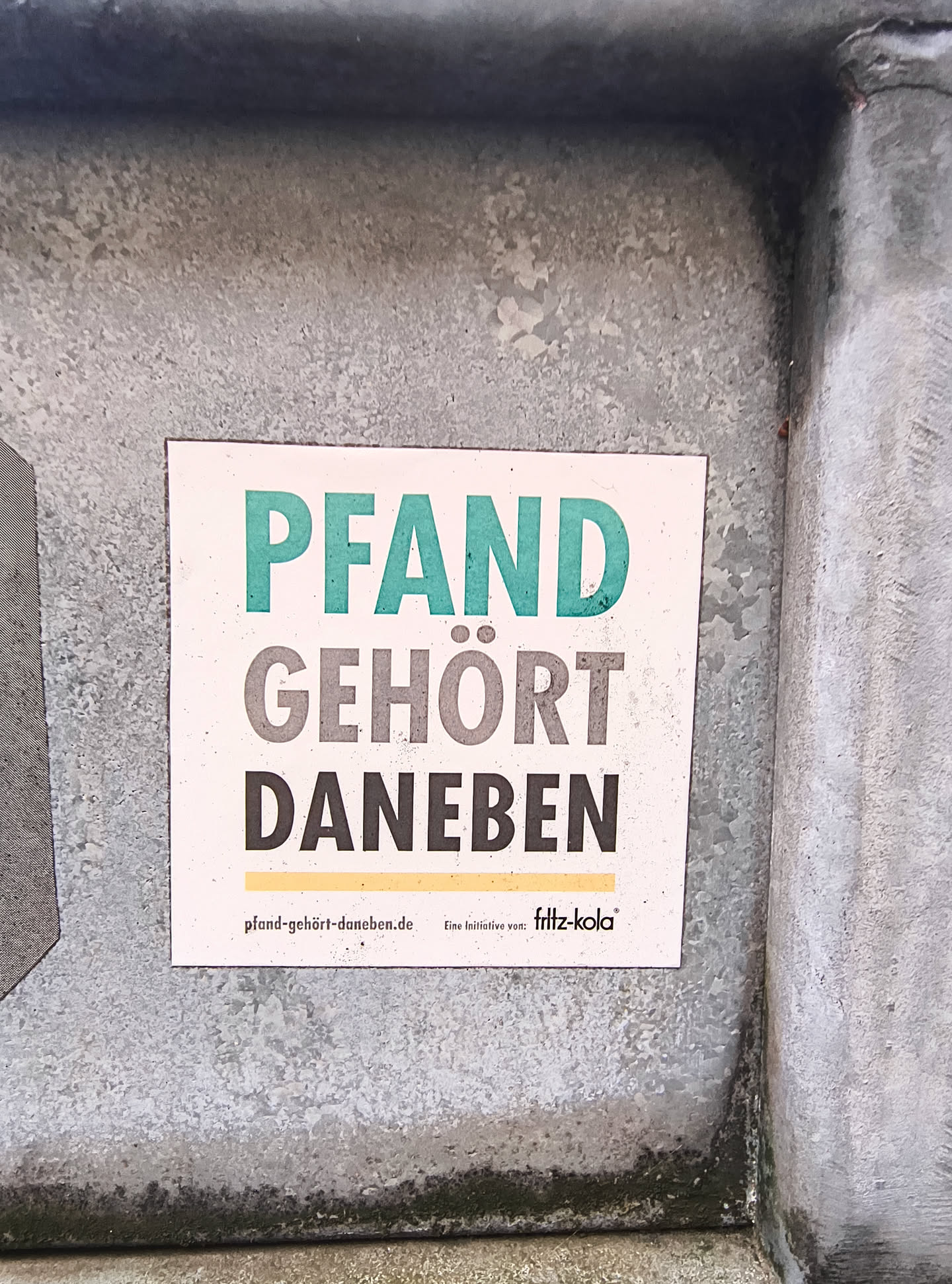 The sticker is square-shaped with a white background. It features large, bold text in the center. The top line reads 'PFAND' in a teal color. The second line reads 'GEHÖRT' in a dark gray color. The third line reads 'DANEBEN' in a dark gray color. Below the main text, there is a thin yellow line. At the bottom left, there is a website address 'pfand-gehört-daneben.de'. To the right of the website, it says 'Eine Initiative von:' followed by the 'fritz-kola' logo in white with a black outline.