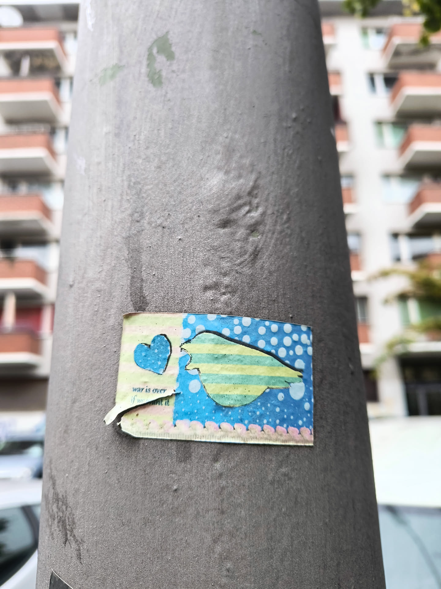 The sticker is rectangular with slightly torn edges, adhered to a grey pole. It features a stylized blue heart on the left side, with a cut-out silhouette of a dove in flight on the right. The dove is depicted with horizontal stripes of light green and yellow, and its body is outlined in blue. The background of the sticker is a vibrant blue with scattered white polka dots. Below the heart, there is some text. The overall aesthetic is handmade and colorful, conveying a message of peace.
