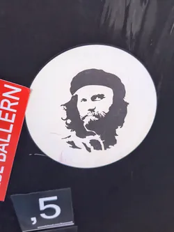 A round white sticker with a black stencil portrait of Che Guevara. He is wearing his iconic beret, and the stencil shows his long hair and beard.