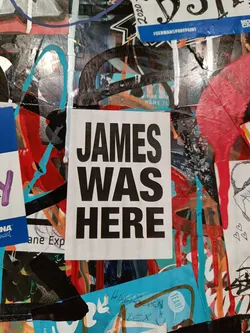 A black and white sticker with the text "JAMES WAS HERE" in bold, uppercase letters. The sticker is affixed to a wall covered in various other stickers and graffiti.
