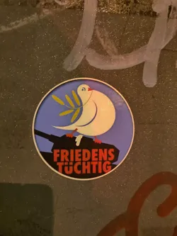 Street sticker A round sticker featuring a white dove carrying an olive branch, perched atop a tank. The text below reads "FRIEDENS TÜHTIG" which translates from German to "Peace-capable". The design promotes peace and disarmament.