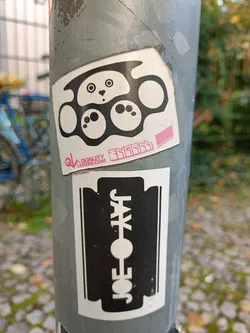 Street sticker A black and white sticker featuring a cute panda face incorporated into a brass knuckles design.  The sticker also has some text that says "SNUGGLE" and a website address, qualitylovers.com. Below it is another sticker of a razor blade with "JOE JAY" written on it. 