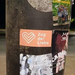 A peach-colored sticker with a double heart symbol and the text "Zug der Liebe" (Train of Love) in white.  The sticker is affixed to a dark-colored pole.