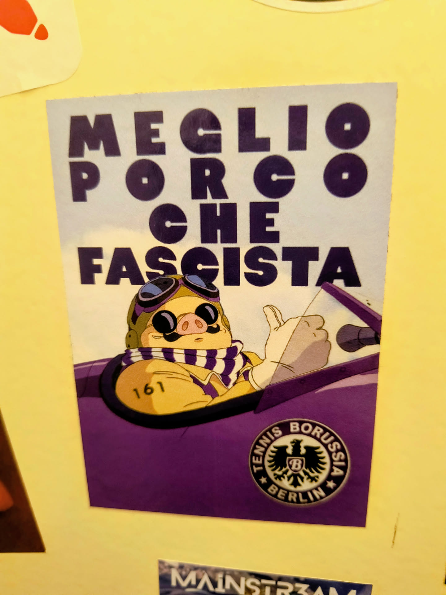 Street sticker The sticker features a cartoon pig character, reminiscent of Porco Rosso, depicted from the chest up, wearing aviator goggles with purple lenses, a brown pilot's helmet, and a white and purple striped scarf. The pig has a prominent mustache and is shown giving a thumbs-up with a white-gloved hand, seemingly driving a purple open-top car. On the pig's left arm, the number '161' is visible. The top portion of the sticker has large, bold, dark purple text against a lighter, almost white background, reading 'MEGLIO PORCO CHE FASCISTA'. In the bottom right corner, there's a circular black and white logo for 'TENNIS BORUSSIA BERLIN' featuring an eagle crest with a 'B' in the center. The overall color scheme is dominated by purple, white, and yellow/tan.