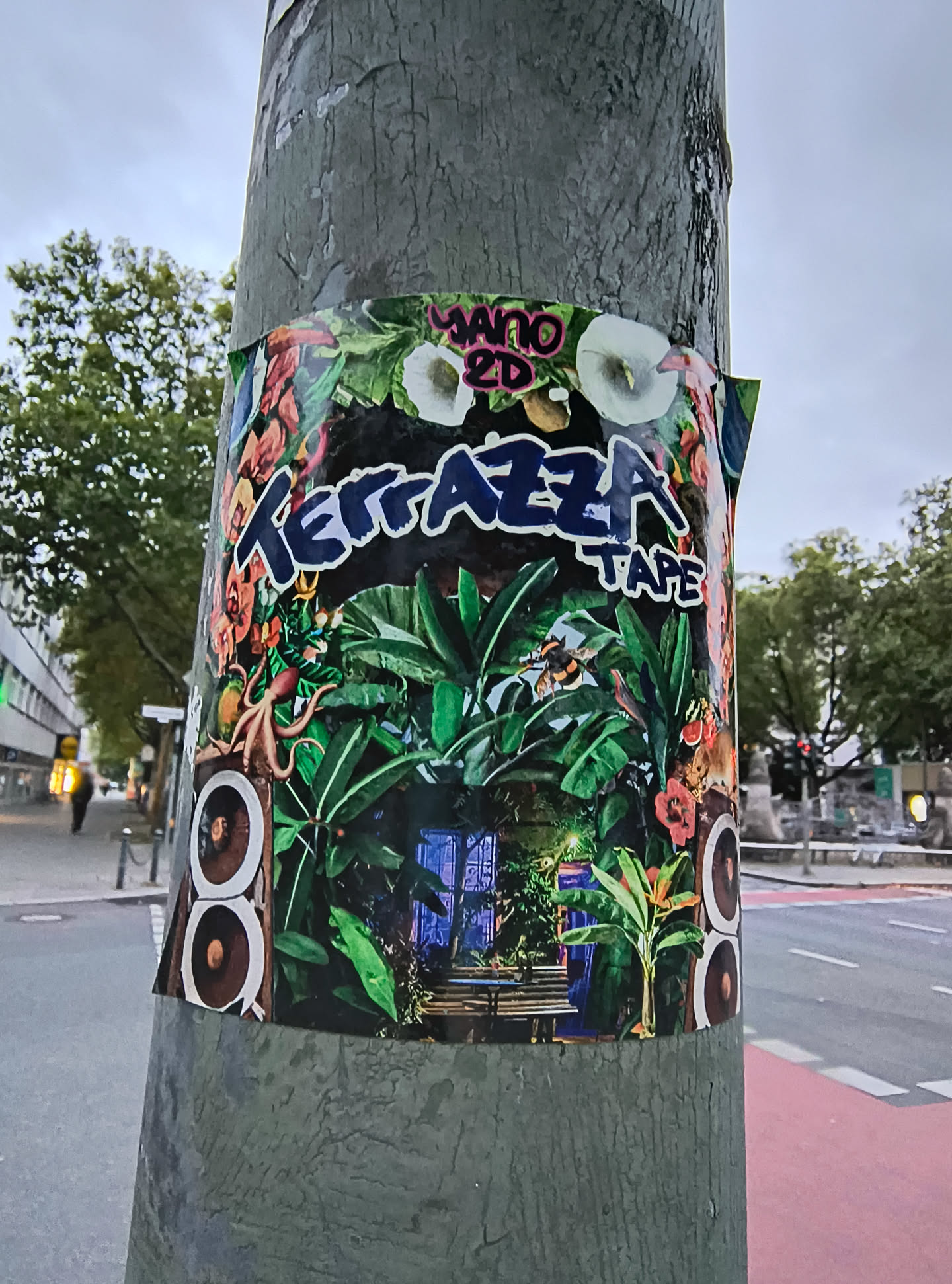 The sticker is a vibrant and colorful collage centered on a pole. It features a lush, tropical jungle scene with dense green foliage, exotic flowers in shades of pink and orange, and white calla lilies. In the foreground, a stylized octopus and a bee are visible. Two sets of stacked circular shapes, resembling speakers, are placed on either side of the central image. The background of the jungle scene appears to be a dimly lit, possibly blue-toned, doorway or entrance with benches. The text 'TERRAZZA TAPE' is prominently displayed in bold, blue, stylized lettering across the top of the sticker, with 'TAPE' in smaller white lettering below. Above 'TERRAZZA', the text 'MANO 2D' is visible in pink and green lettering. The overall aesthetic is a blend of nature, music, and a slightly surreal or psychedelic feel.