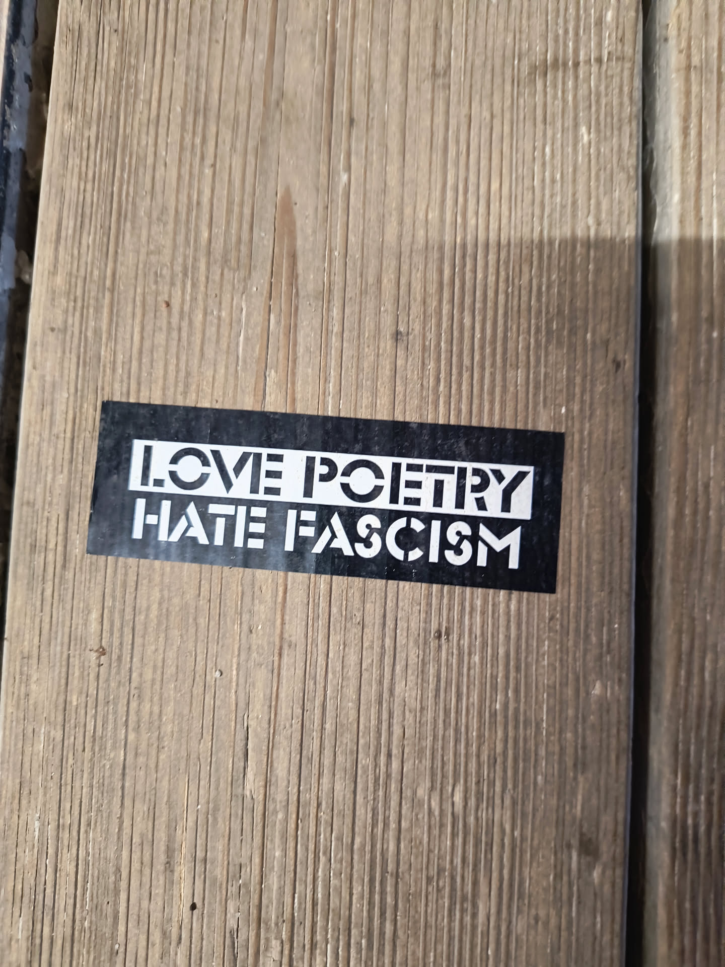 The sticker is rectangular with a black background and white text. The text is arranged in two lines. The top line reads 'LOVE POETRY' and the bottom line reads 'HATE FASCISM'. The font is sans-serif with a stencil-like appearance, where some letters have cutouts. The sticker appears to be adhered to a wooden surface.