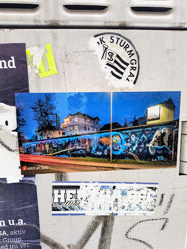 A sticker featuring a picture of a colourful mural at night. The mural seems to represent global issues or interconnectedness. There's a YouTube link visible at the bottom of the sticker image.  Other stickers are visible on the surface, including one with partially visible text that looks like a logo or abbreviation.