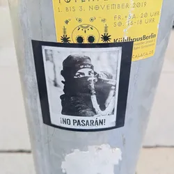 Street sticker A black and white photo sticker showing a woman wearing a balaclava aiming a rifle or similar weapon. The text "¡NO PASARÁN!" is written at the bottom. The sticker appears to be wheat-pasted on a metal pole.