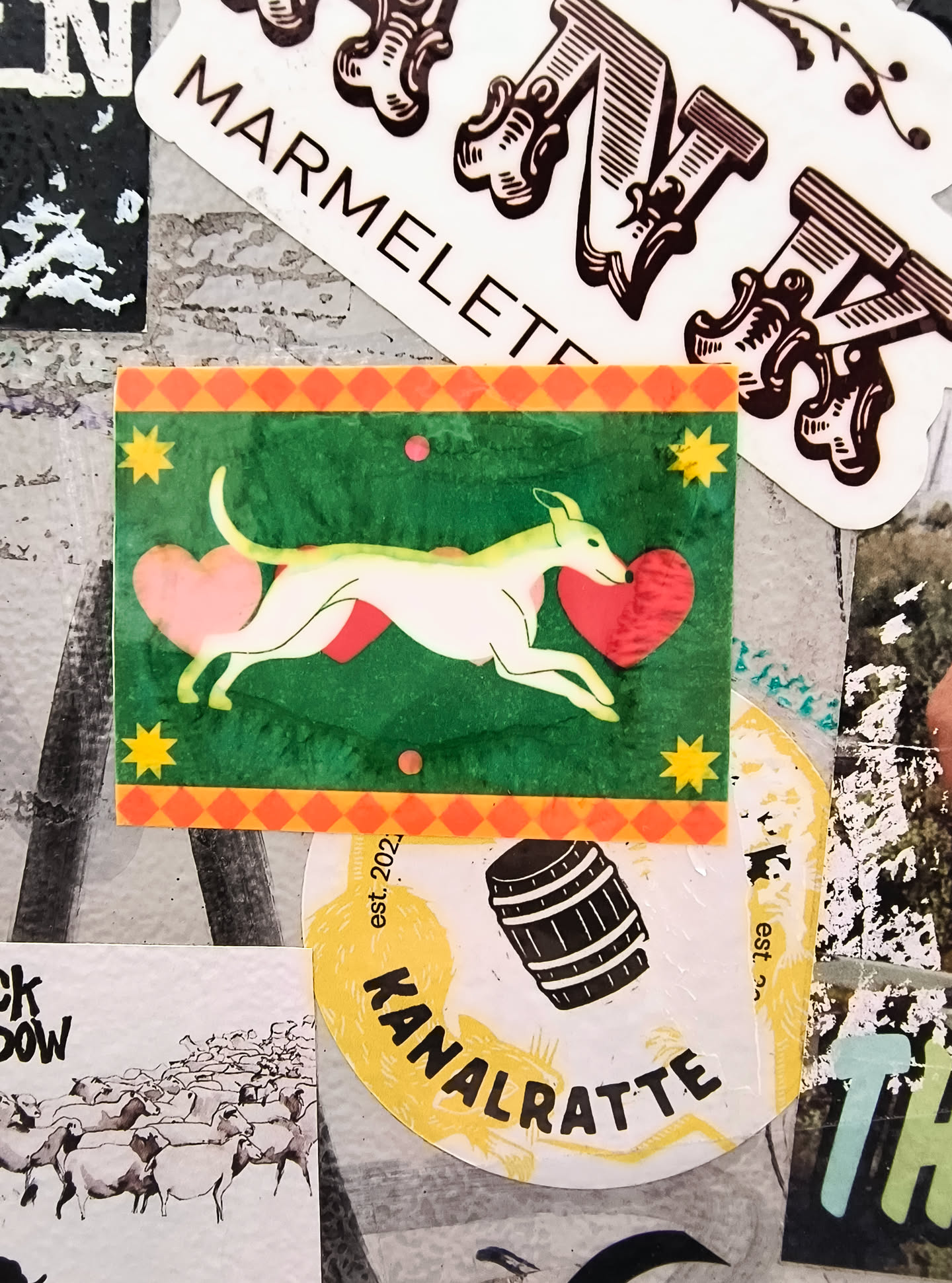 The sticker features a stylized, light-colored whippet in mid-stride, with its body arcing. It is depicted against a rich green background. Two pink hearts are prominent: one is held in the dog's mouth, and another is positioned near its hindquarters. The top and bottom edges of the sticker are bordered by a pattern of orange diamonds. Scattered across the green background are four yellow starbursts and two small pink circles. The overall aesthetic is whimsical and charming.