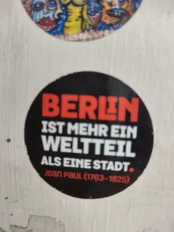 A black and red sticker with a quote by Jean Paul: "Berlin is more a part of the world than a city."  The sticker is round and has white text on a black background. The quote is in German.