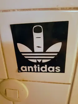 Street sticker A rectangular black sticker featuring a white graphic that parodies the iconic Adidas trefoil logo. The upper part of the trefoil is replaced by a stylized white middle finger gesture, while the lower three horizontal stripes remain. Below this altered logo, the word 'antidas' is written in white, sans-serif capital letters. The sticker is applied to a light-colored tiled surface and shows minor creasing at the bottom left corner, indicating its physical nature.