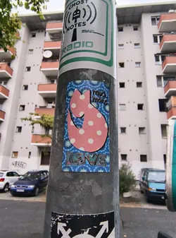 Street sticker The sticker is predominantly blue with a repeating wave-like pattern. Centered on the sticker is a stylized pink cat silhouette filled with white polka dots. The cat has a curved tail and appears to be in a playful pose. Above the cat, in a darker pink, are the words 'much more'. Below the cat, in a colorful, graffiti-style font with pink, blue, and green hues, is the word 'LOVE'. The overall aesthetic is cheerful and whimsical.
