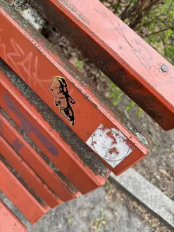 Street sticker The sticker features a stylized, somewhat plump baby or cherub-like figure, rendered with a golden-yellow fill and distinct black outlines. The figure is depicted in a dynamic pose, appearing to walk or run, with its arms slightly bent and legs apart. Its head is proportionally large, and the facial features are simplified. The sticker has a slightly glossy finish and is affixed to a heavily weathered, orange-painted wooden slat of a bench, which shows significant signs of wear, dirt, and possibly lichen. To the right of this sticker, there is a partially removed, circular white sticker with faded reddish markings, which is not the primary focus.