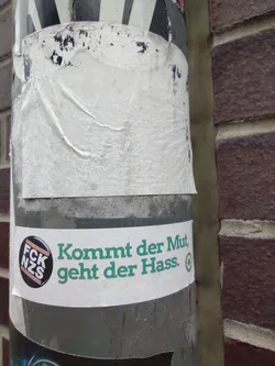 A sticker with the text "Kommt der Mut, geht der Hass."  which translates to "When courage comes, hate goes." in German. It features a smaller circular sticker with the abbreviation "FCK NZS".