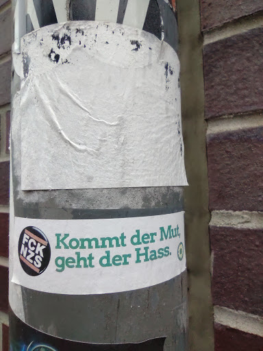 Street sticker A sticker with the text "Kommt der Mut, geht der Hass."  which translates to "When courage comes, hate goes." in German. It features a smaller circular sticker with the abbreviation "FCK NZS".