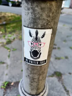 A sticker featuring a zebra with pink glasses and the phrase 'We prefer free'. The sticker is affixed to a weathered gray pole.