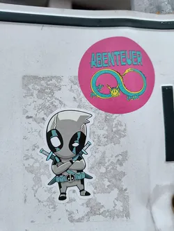 A sticker featuring a chibi-style grey Deadpool with crossed katanas.  Another sticker in the background says "Abenteuer" (German for Adventure) with a stylized infinity symbol and a smiley face. 