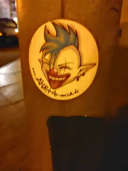 Street sticker Round sticker featuring a cartoon illustration of a clown with blue spiky hair, a red nose, and a wide, toothy grin. The clown's tongue is sticking out.  The text 'alry-ch-musik.de' is visible on the sticker.