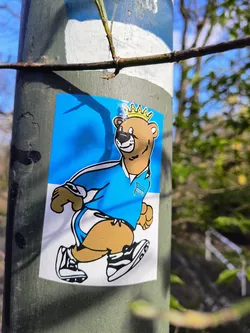 Street sticker A sticker featuring a cartoon illustration of a bear wearing a crown and a blue shirt while running. The bear is depicted in a dynamic pose, suggesting movement and action. The color scheme is simple, using mostly blue and brown. The sticker is applied to a gray pole in an outdoor setting.