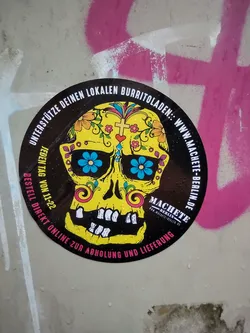Sticker featuring a yellow sugar skull with floral details. The text around the skull says to support your local burrito shop and includes a website address.  The text also mentions ordering online for pickup and delivery.