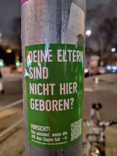 A green sticker posted on a pole. The text on the sticker is in German and questions whether someone's parents were born locally. It warns that this is what happens when the AfD (Alternative for Germany) has a say. There's also a QR code linking to a website.