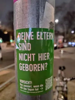 Street sticker A green sticker posted on a pole. The text on the sticker is in German and questions whether someone's parents were born locally. It warns that this is what happens when the AfD (Alternative for Germany) has a say. There's also a QR code linking to a website.