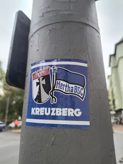 Street sticker The sticker is predominantly blue with white and black accents. It features a crest on the left side, which includes a red-topped castle-like structure with a black cross superimposed on a brick pattern. Above the cross, a small bear is visible. To the right of the crest, a blue flag with a white outline waves, displaying the text 'Hertha BSC' in white, stylized font. Below the crest and flag, the word 'KREUZBERG' is written in large white capital letters, separated by a thin white horizontal line from the upper design.