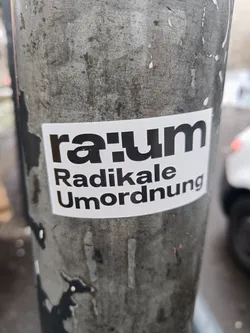A white sticker with black text on a gray pole. The main text is "raum" in a bold, stylized font, and below it are the words "Radikale Umordnung".  The sticker appears to be weathered, showing some signs of wear.