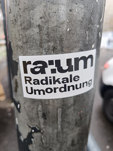 Street sticker A white sticker with black text on a gray pole. The main text is "raum" in a bold, stylized font, and below it are the words "Radikale Umordnung".  The sticker appears to be weathered, showing some signs of wear.