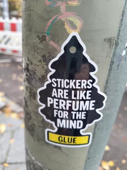 Street sticker The sticker is shaped like a classic car air freshener, specifically the pine tree silhouette. It is predominantly black with a white outline. White text is printed on the black surface, reading 'STICKERS ARE LIKE PERFUME FOR THE MIND'. Below this, there is a bright yellow rectangular bar with the word 'GLUE' in black capital letters. The sticker has a small hole at the top, suggesting it could be hung.
