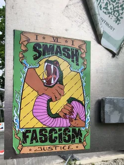 The sticker is a rectangular, vertically oriented design with a vibrant green border featuring stylized blue flame-like accents on the sides. The top and bottom sections are framed by brown, scroll-like banners. The central image is set against a bright yellow background with diagonal black lines. It depicts two brown-skinned hands with dark red fingernails firmly gripping and choking a green snake with a pink, segmented underside. The snake's head is shown with its mouth wide open, revealing sharp white fangs and a red tongue. A small, stylized swastika symbol is subtly integrated into the snake's head pattern. The word "SMASH" is prominently displayed in large, cracked black letters on the top brown banner. Below the central illustration, on the bottom brown banner, the word "FASCISM" appears in similar cracked black lettering. Beneath "FASCISM", the word "JUSTICE" is written in smaller, stylized black letters, flanked by two small stars. The overall aesthetic is bold, illustrative, and carries a strong political message.