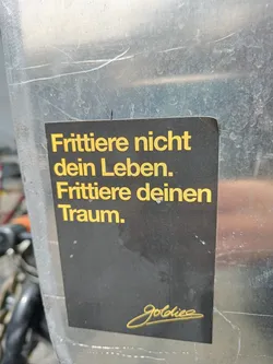 Black and gold sticker with a motivational quote in German. The text translates to "Don't fry your life. Fry your dream."  It's signed with a stylized signature, possibly the artist's name.
