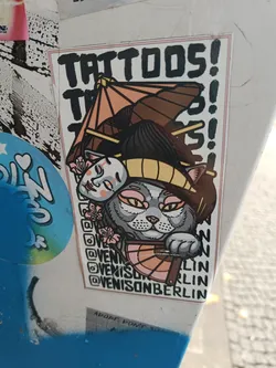 A sticker featuring a cute cat wearing a geisha-inspired hat and holding a hand fan. The design is in a vibrant color palette and has a unique blend of Japanese and modern aesthetics. The sticker also displays the artist's Instagram handle, @venisonberlin, repeated several times. The text 'TATTOOS!' is prominently displayed at the top.