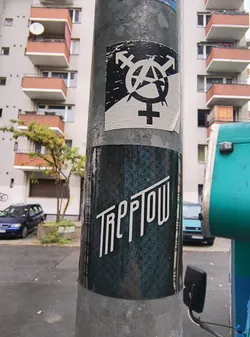 Street sticker The sticker at the center of the image features a black and white design. The top portion displays a combination of symbols: the anarchy symbol (a circle with an 'A' inside) is overlaid with the transgender symbol (an arrow pointing up and to the right, combined with a circle and a cross). Below this, the Venus symbol (a circle with a cross) is prominently featured. The sticker appears to be slightly worn and peeling at the edges, indicating it has been affixed to the pole for some time. The background of the sticker is white, with the symbols rendered in black. Below this sticker, another sticker is visible, predominantly dark teal with a repeating geometric pattern. This lower sticker has white text in a stylized font that reads 'TREPTOW'.