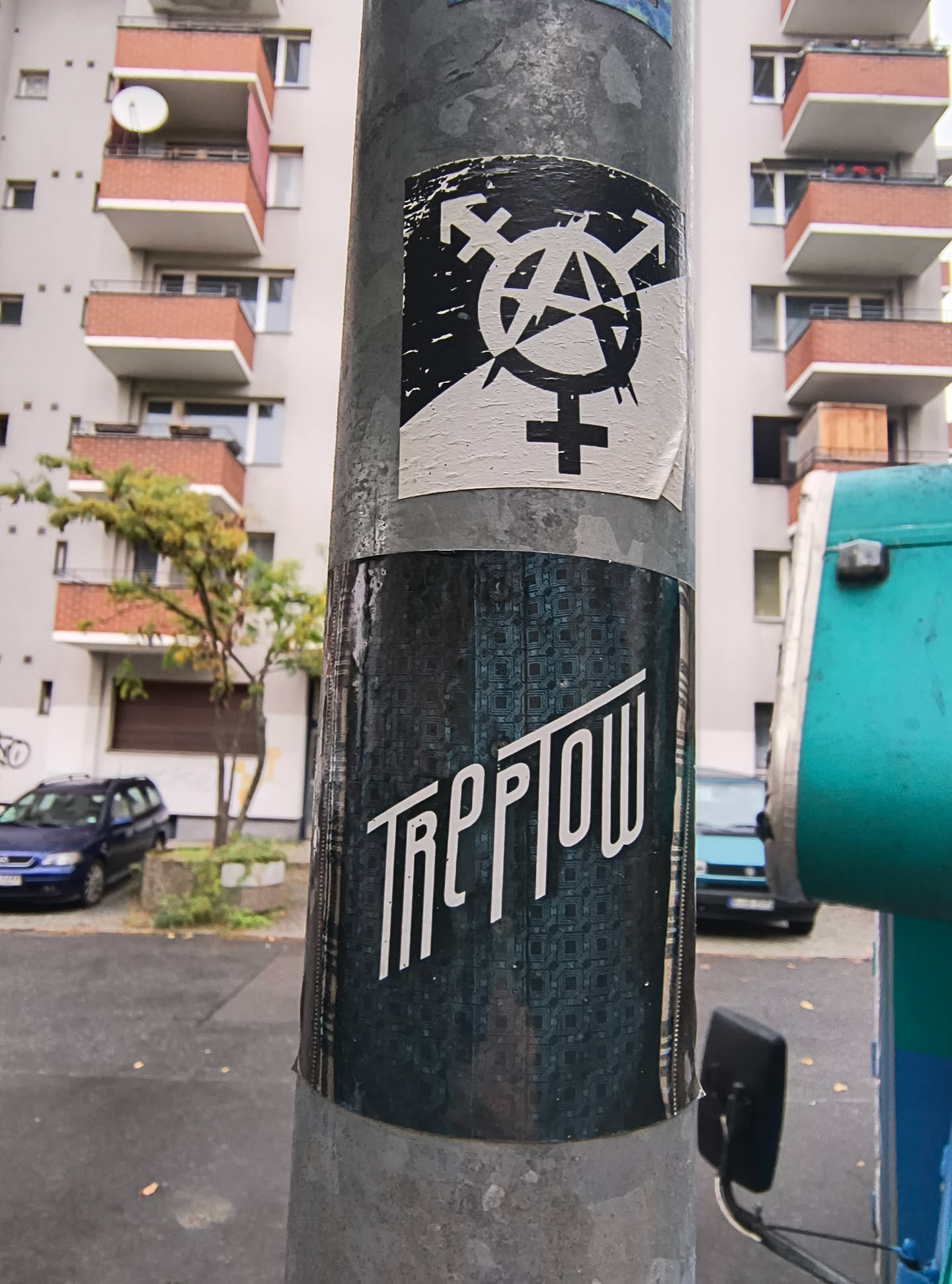 The sticker at the center of the image features a black and white design. The top portion displays a combination of symbols: the anarchy symbol (a circle with an 'A' inside) is overlaid with the transgender symbol (an arrow pointing up and to the right, combined with a circle and a cross). Below this, the Venus symbol (a circle with a cross) is prominently featured. The sticker appears to be slightly worn and peeling at the edges, indicating it has been affixed to the pole for some time. The background of the sticker is white, with the symbols rendered in black. Below this sticker, another sticker is visible, predominantly dark teal with a repeating geometric pattern. This lower sticker has white text in a stylized font that reads 'TREPTOW'.
