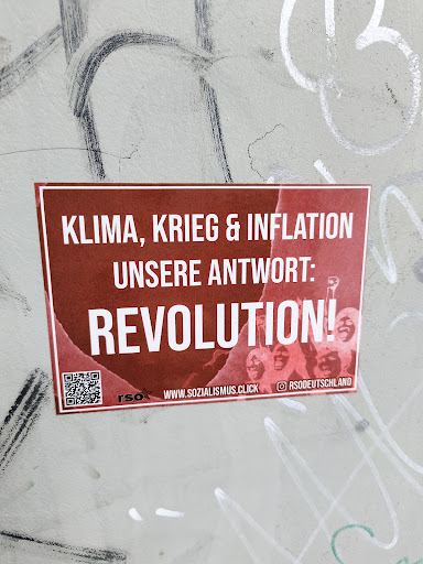 A red sticker with white text pasted on a wall. The text reads "Klima, Krieg & Inflation, Unsere Antwort: Revolution!". It also contains a QR code and the social media link to RSOdeutschland's Instagram page.