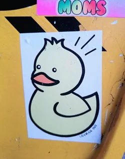 A simple sticker featuring a cartoon drawing of a cute yellow duck. The duck has a simplistic design with a few lines indicating movement or excitement. The artist's signature, LOVMAN.ONE, is visible at the bottom.