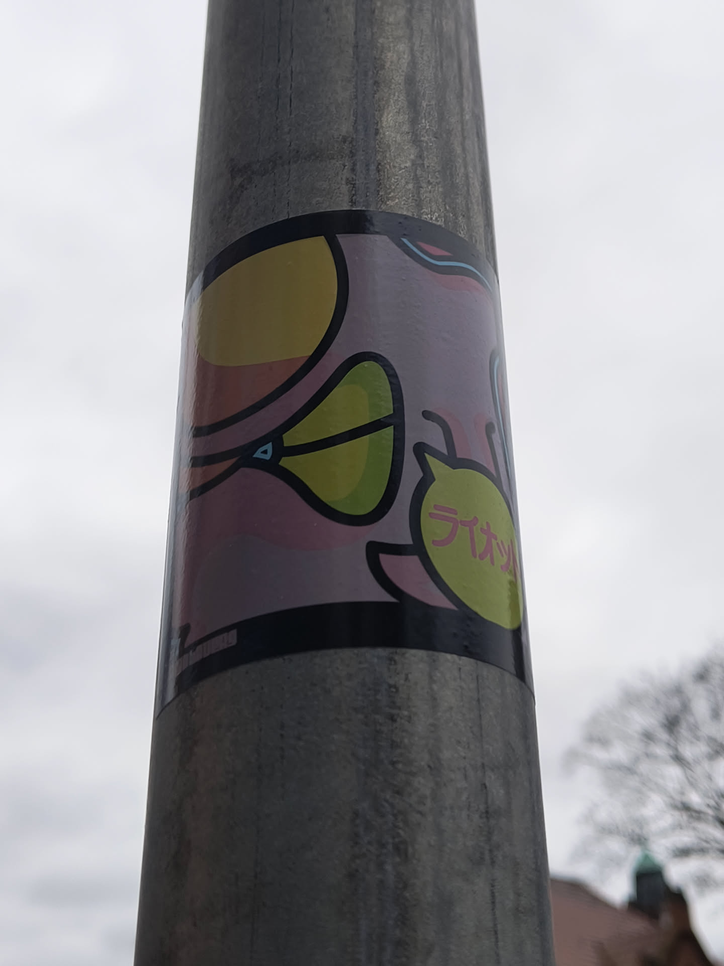 The sticker is rectangular and applied vertically on a textured metal pole. Its design is characterized by thick black outlines enclosing abstract, cartoonish shapes. The background of the sticker features a soft gradient of light pink and purple hues. On the left, there's a large, irregular yellow shape, possibly an eye or a stylized fruit, connected to a smaller, lime-green, leaf-like form with a tiny blue triangular accent. To the right, a prominent lime-green, rounded shape, resembling a speech bubble or another fruit, contains pink Japanese characters. The overall aesthetic is playful and graphic, utilizing a distinct color palette of yellow, lime green, pink, purple, and black outlines. A small, unreadable signature or logo is faintly visible at the bottom left edge.
