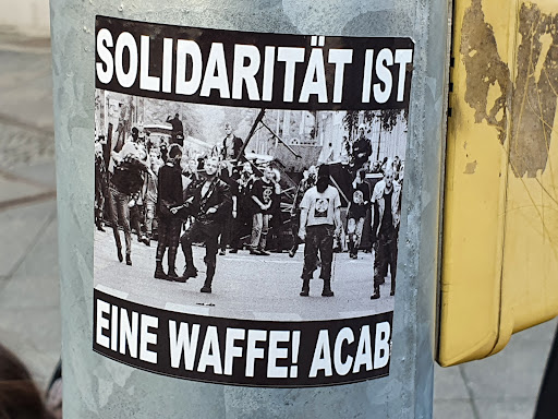 Street sticker Black and white photo of a protest or riot with text overlay. The text on the sticker translates to: 'Solidarity is a weapon! ACAB'