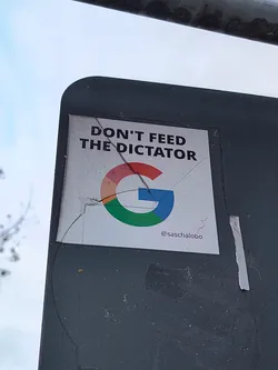 Street sticker The sticker is rectangular with a white background. At the top, in bold black sans-serif font, it reads 'DON'T FEED THE DICTATOR'. Below this text is a stylized Google 'G' logo, with its characteristic red, yellow, green, and blue segments. The sticker appears to be slightly creased and cracked, suggesting it has been applied outdoors. At the bottom right of the sticker, a social media handle '@saschalobo' is visible in smaller black text.